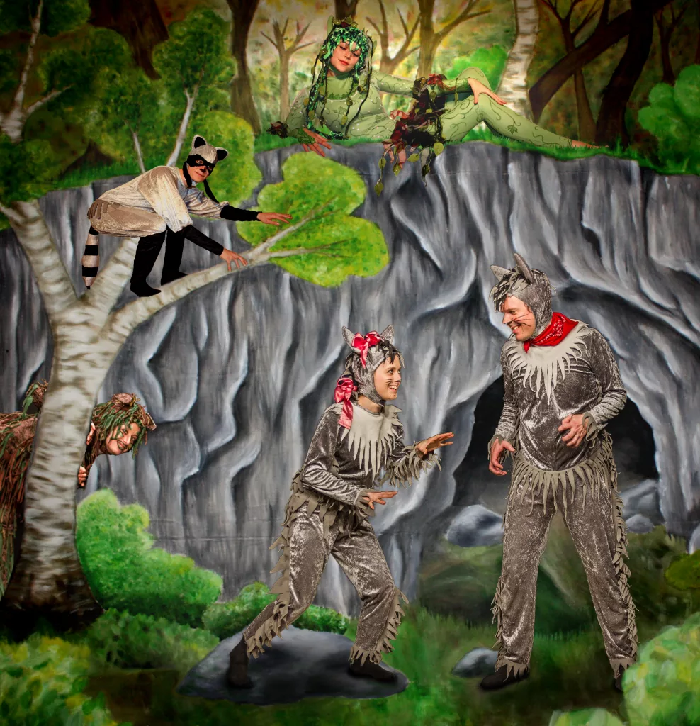 Das Plakat des Theaterstücks "Wolfswege". Die fünf Schauspielenden stehen vor einer gemalten Höhle im Wald. Sie tragen Tierkostüme: ein Waschbär, ein Stinktier, Robbe, eine Waldfee und zwei Wölfe.